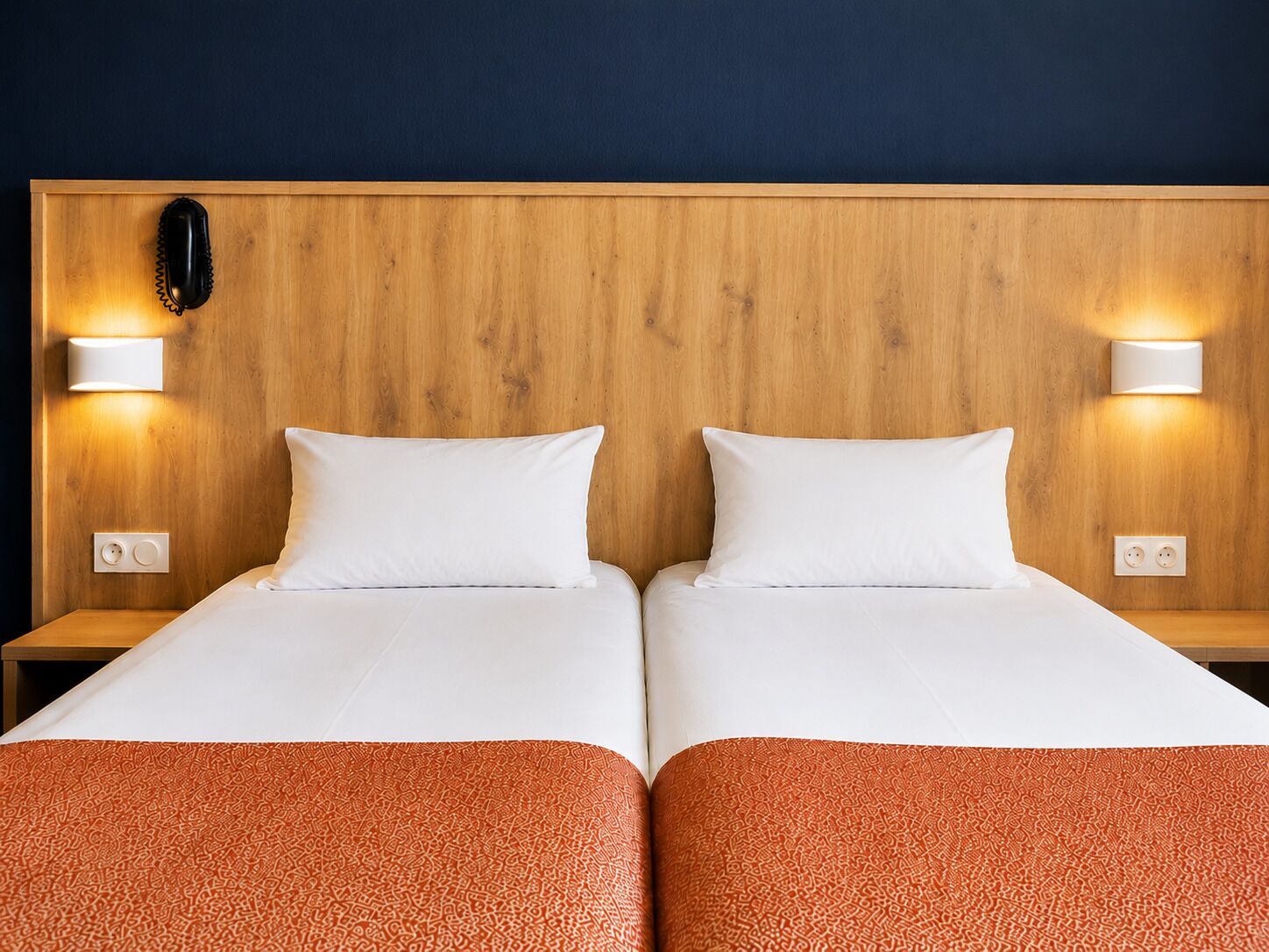 Une chambre d’hôtel avec deux lits, une tête de lit en bois, des appliques murales et un téléphone.