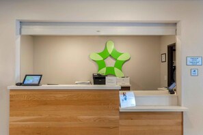 A hotel reception desk with a wooden counter, a digital screen, a printer, and a sign indicating check-in and check-out times. - Extended Stay America Select Suites - Charlotte - Northlake (Charlotte)