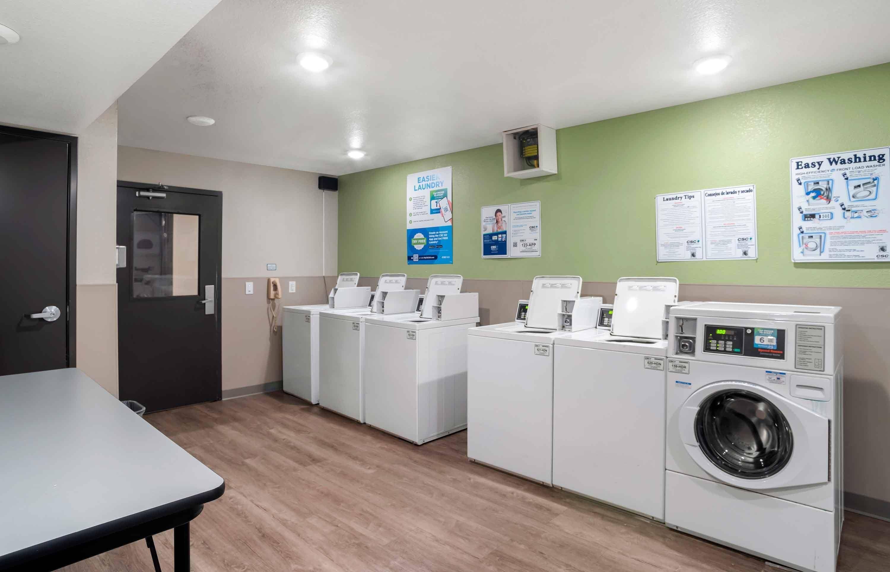 A laundry facility with multiple washing machines and dryers, a table, and informational posters on the wall.