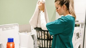 Eine Person in türkisfarbenem OP-Kleid hängt ein weißes Handtuch in einen Wäschekorb auf eine weiße Waschmaschine.