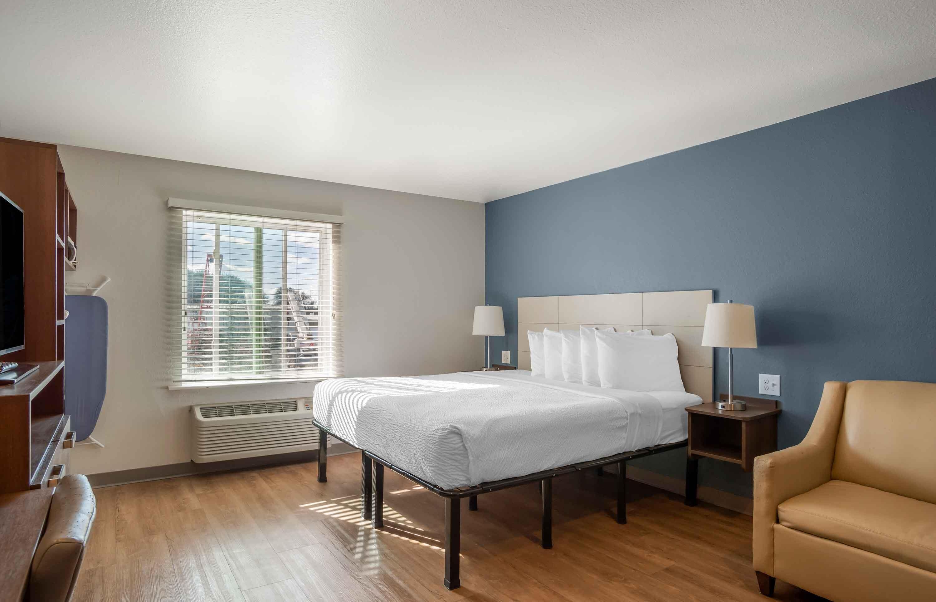 Ein Hotelzimmer mit Bett, Nachttischlampen, einem Sessel, einem Fernseher und einem Fenster mit Jalousien.