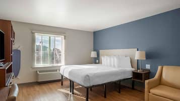 Ein Hotelzimmer mit Bett, Nachttischlampen, einem Sessel, einem Fernseher und einem Fenster mit Jalousien.