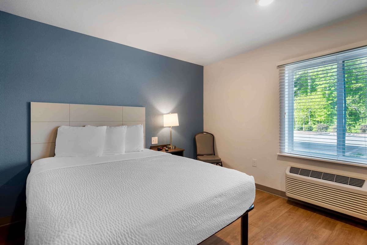 A hotel room with a bed, a nightstand, a lamp, a chair, and a window with blinds.