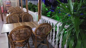 Ein Restaurant mit Korbstühlen und einem Holztisch, umgeben von einer weißen Balustrade und Grünpflanzen.