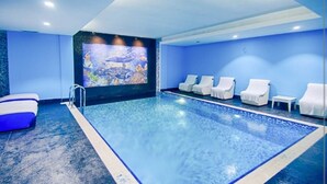 Una piscina interior moderna con suelo de baldosas azules, un mural en la pared y varias sillas reclinables blancas.