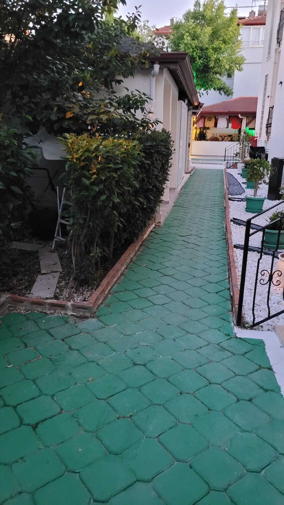 Uma passagem estreita com piso verde que conduz a um edifício com fachada branca e telhado vermelho.
