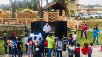 Un grupo de niños reunidos alrededor de un escenario con altavoces, en un entorno al aire libre con una estructura de ladrillo y árboles al fondo.