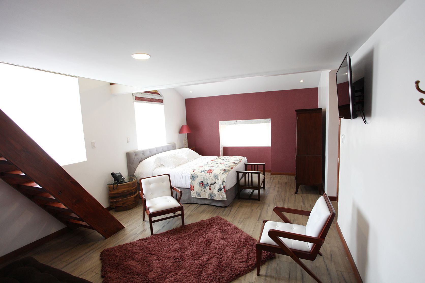 Un dormitorio con cama, dos sillas, televisor, aparador de madera, alfombra roja y ventana.