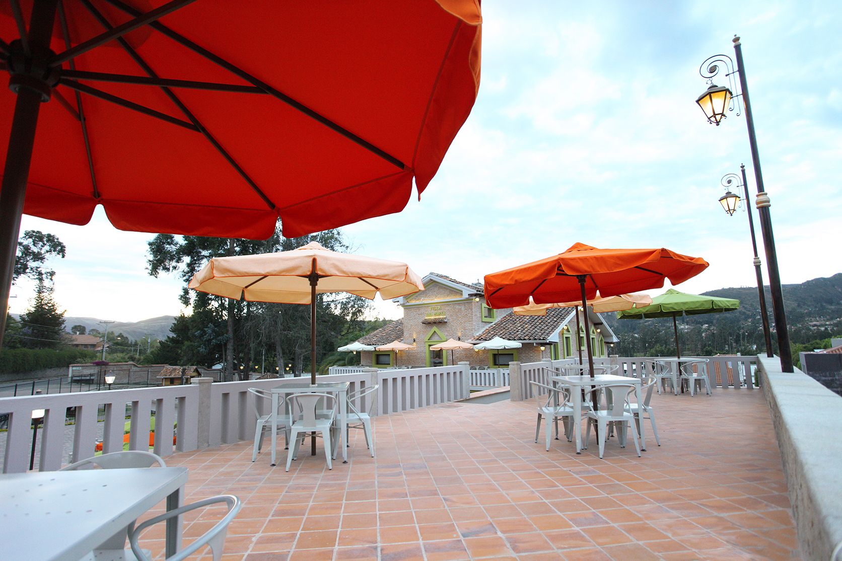 An outdoor patio area with white tables and chairs, orange and green umbrellas, and a brick building in the background.