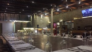 Uma longa mesa de jantar posta com vários lugares, incluindo copos de vinho, talheres e pratos, em restaurante com paredes de pedra e vigas de madeira.