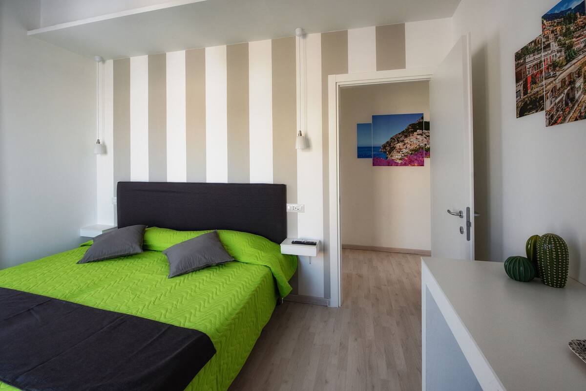 A bedroom with a bed, a bedside table, a nightstand, a door, and a wall with a painting.