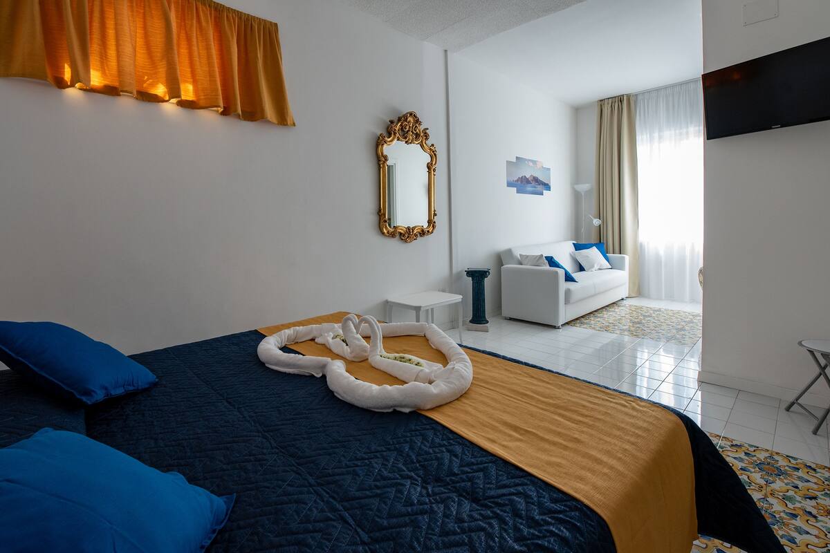 A hotel room with a bed, a white armchair, a small table, and a TV mounted on the wall.