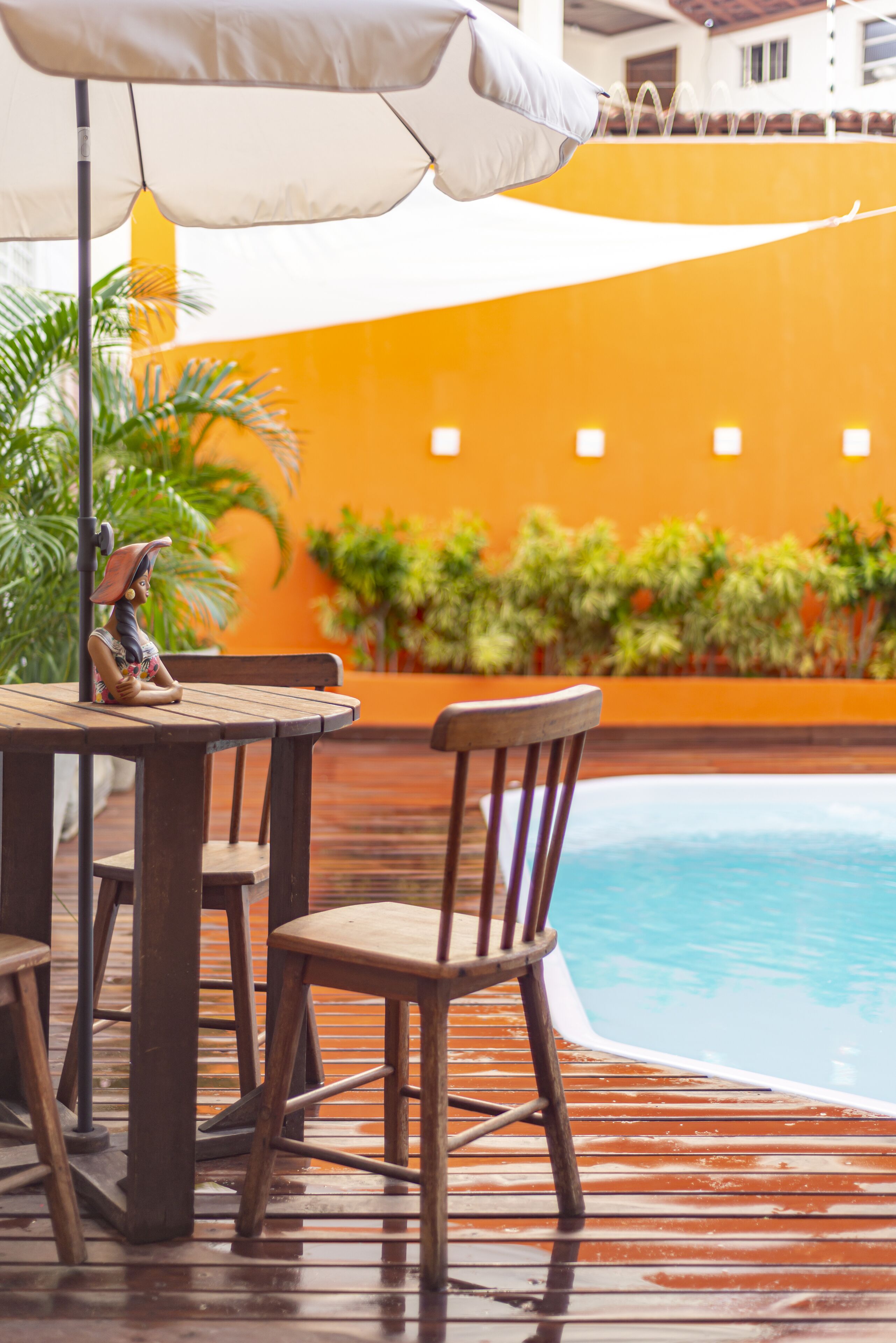 Un área de piscina con madera, una mesa, sillas, una sombrilla blanca y una estatua.