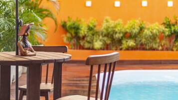 Un área de piscina con madera, una mesa, sillas, una sombrilla blanca y una estatua.
