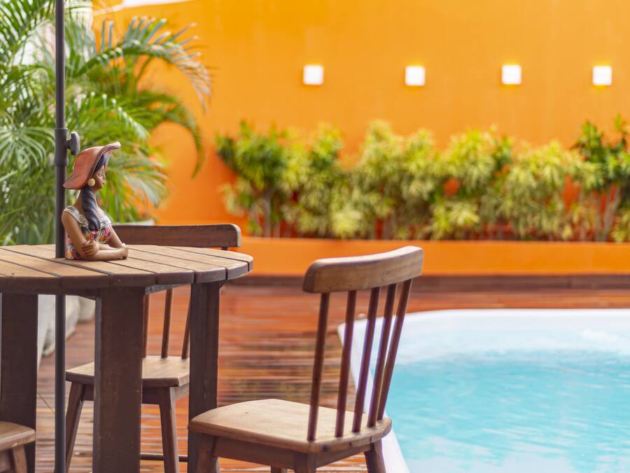Un área de piscina con madera, una mesa, sillas, una sombrilla blanca y una estatua.