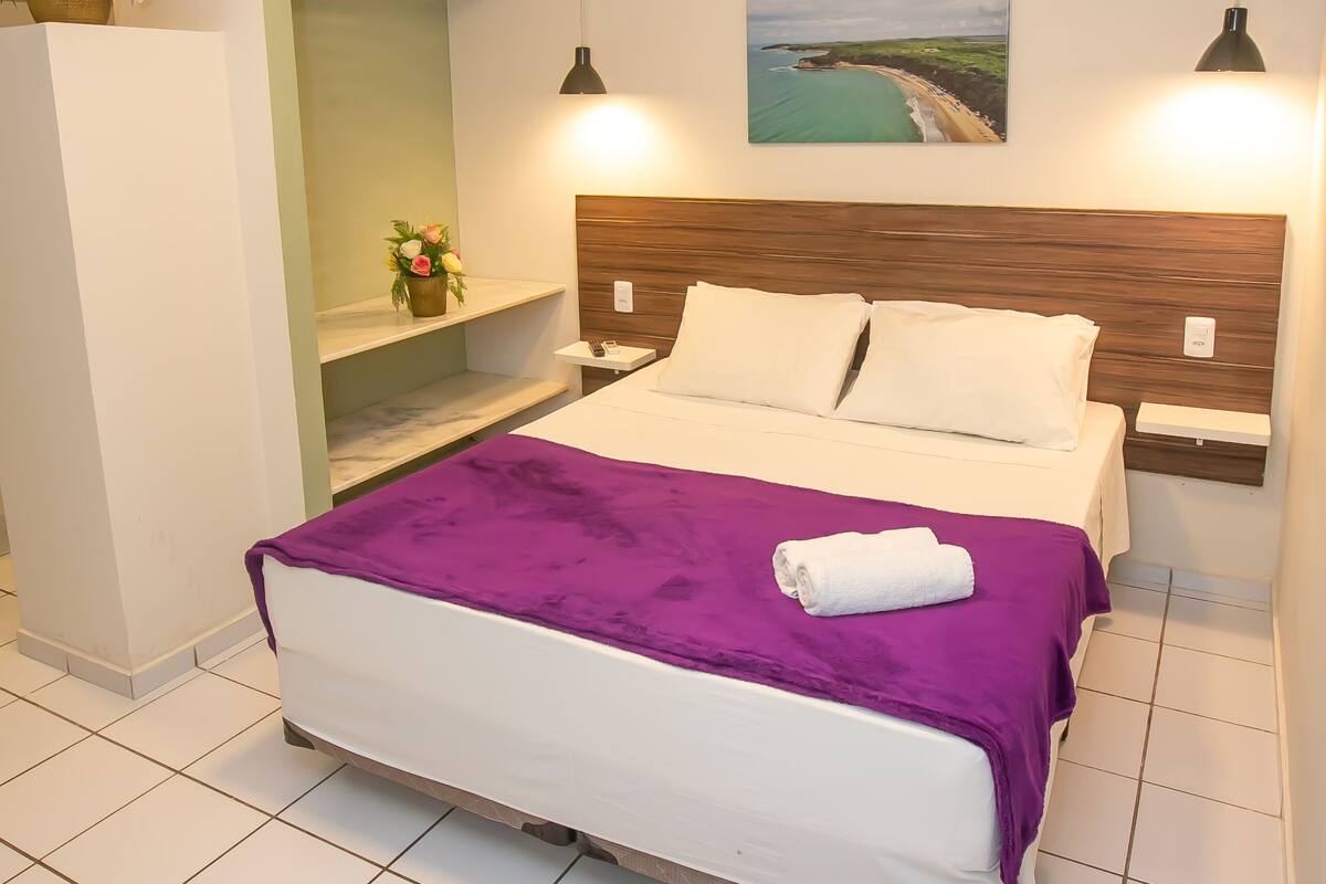 A neatly arranged bedroom with a bed, bedside tables, a nightstand, a wall-mounted shelf, and a painting.