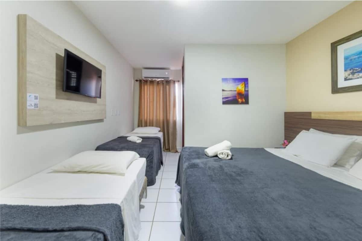 A hotel room with three single beds, a flat-screen TV mounted on the wall, a painting on the wall, and a window with curtains.