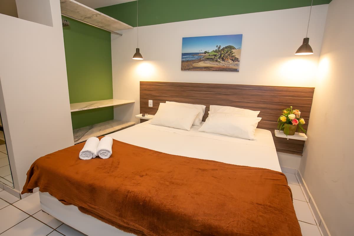 A neatly made bed with a brown blanket and two rolled towels, a wooden headboard, a nightstand with a vase of flowers, and a painting of a beach scene on the wall.