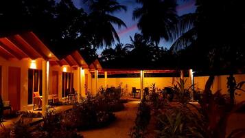 A covered outdoor area with seating and a pathway, surrounded by vegetation and palm trees.