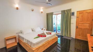 A bedroom with a bed, a ceiling fan, a wooden door, and a view of greenery outside.