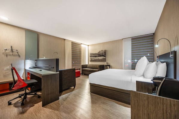 A modern hotel room with a large bed, a desk with a chair, a sofa, and a red chair.
