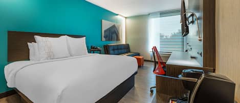 A modern hotel room with a large bed, a desk with a coffee maker, a TV, and a window with blinds.