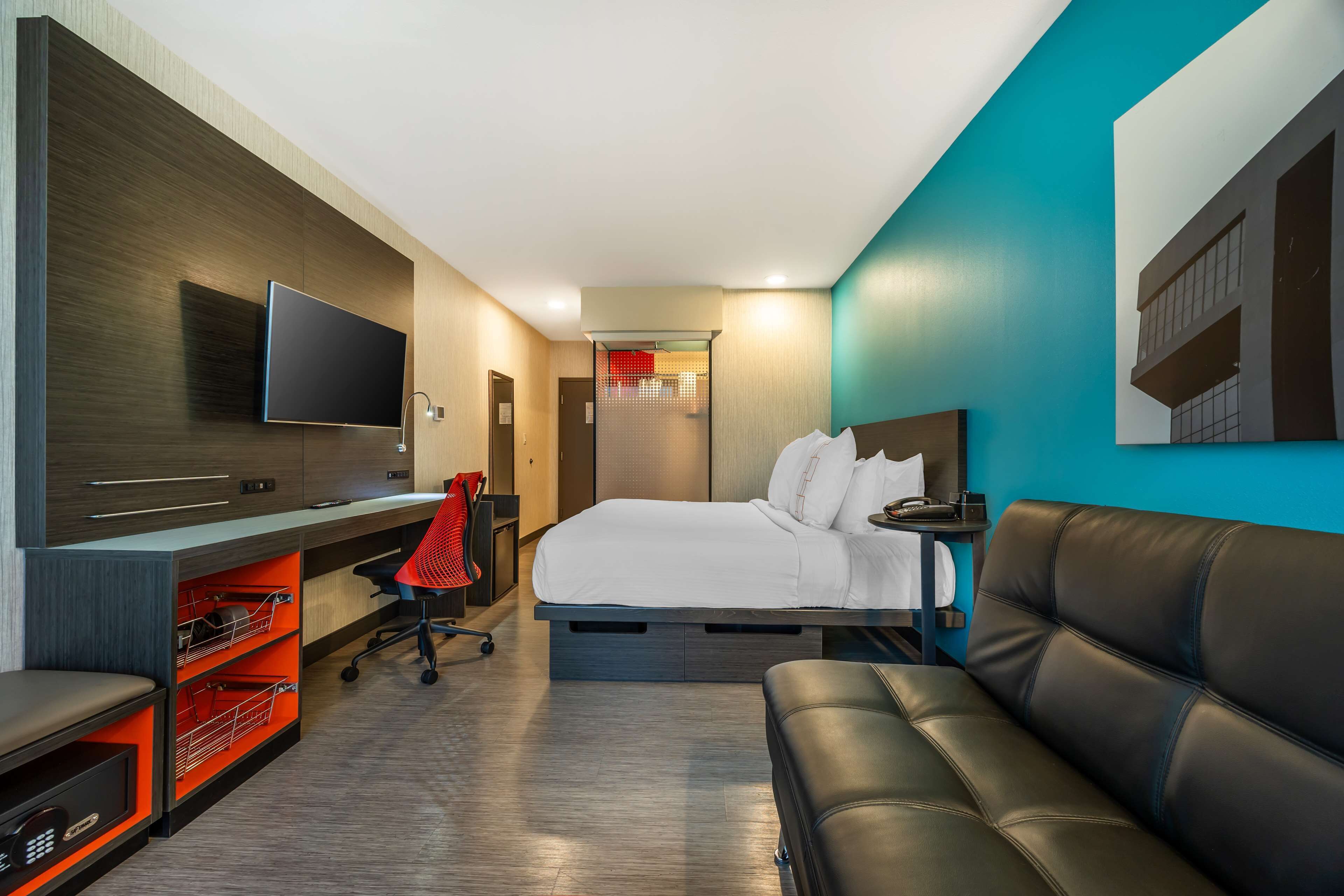 A modern hotel room with a bed, a desk with a flat-screen TV, a leather sofa, and a red chair.