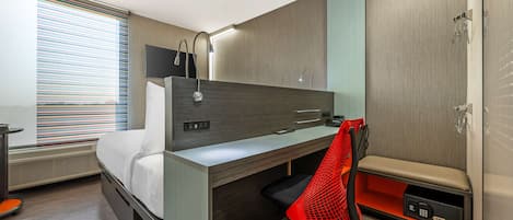 Ein modernes Hotelzimmer mit Holzschreibtisch, einem roten Mesh-Stuhl, einem Bett, einem Fernseher und einem Duschbereich.