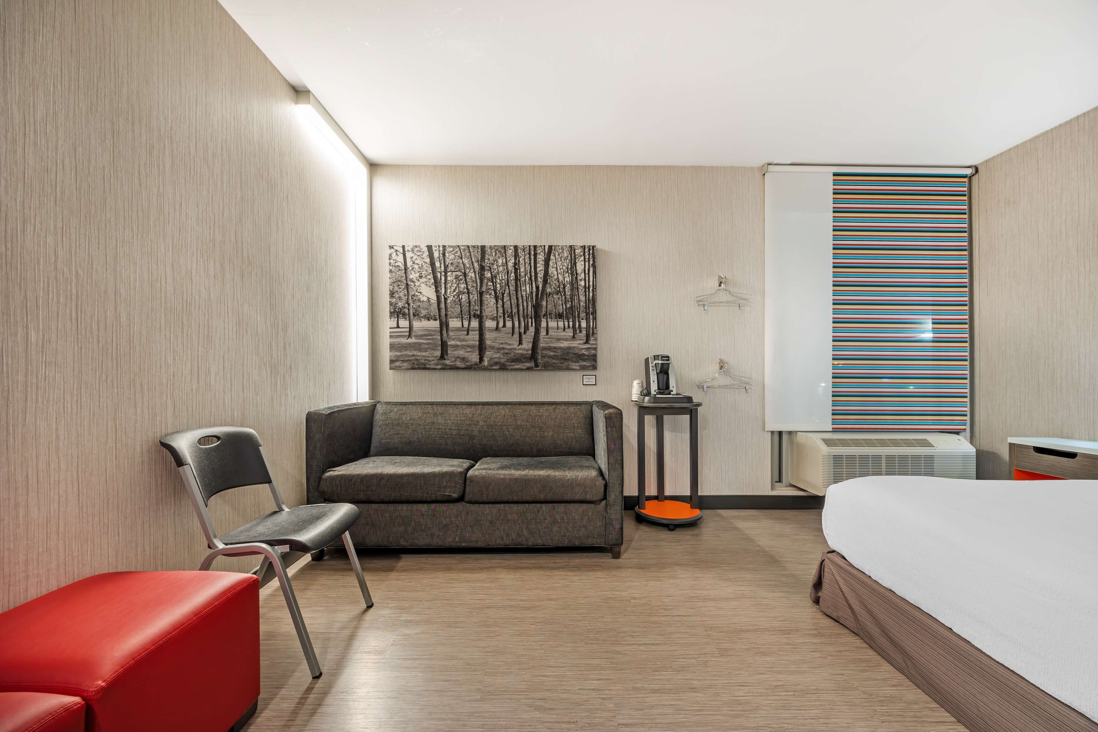 Une chambre d’hôtel avec un canapé gris, un fauteuil rouge, un lit, une petite table avec une lampe et une fenêtre avec des stores à rayures.