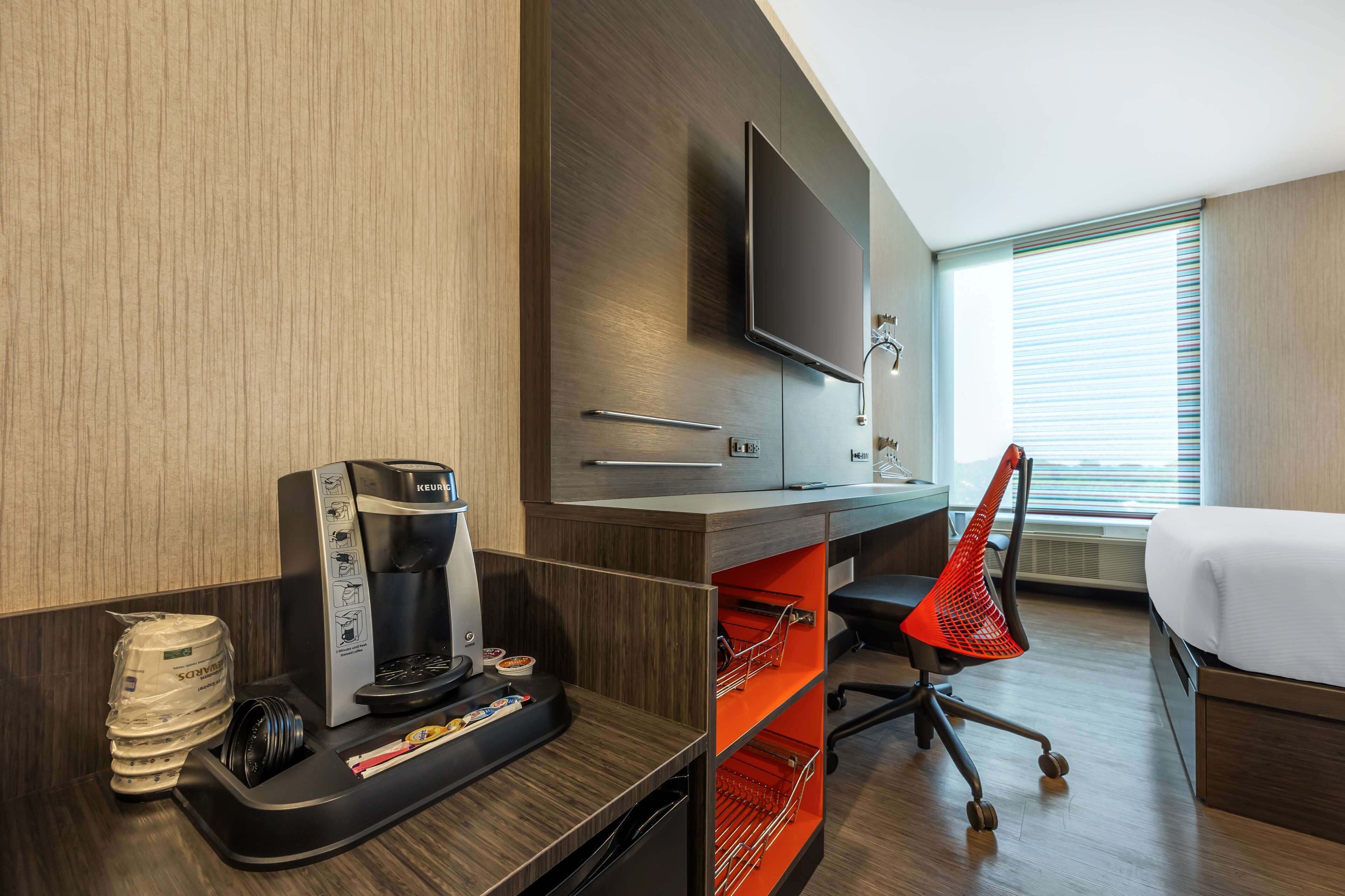 Habitación de hotel con cama, escritorio con cafetera, televisor y ventana con persianas.