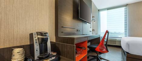Habitación de hotel con cama, escritorio con cafetera, televisor y ventana con persianas.