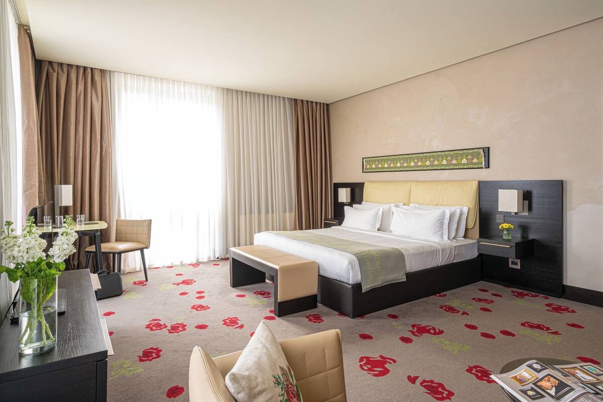 Habitación de hotel con cama, escritorio, silla, banco y un jarrón con flores.