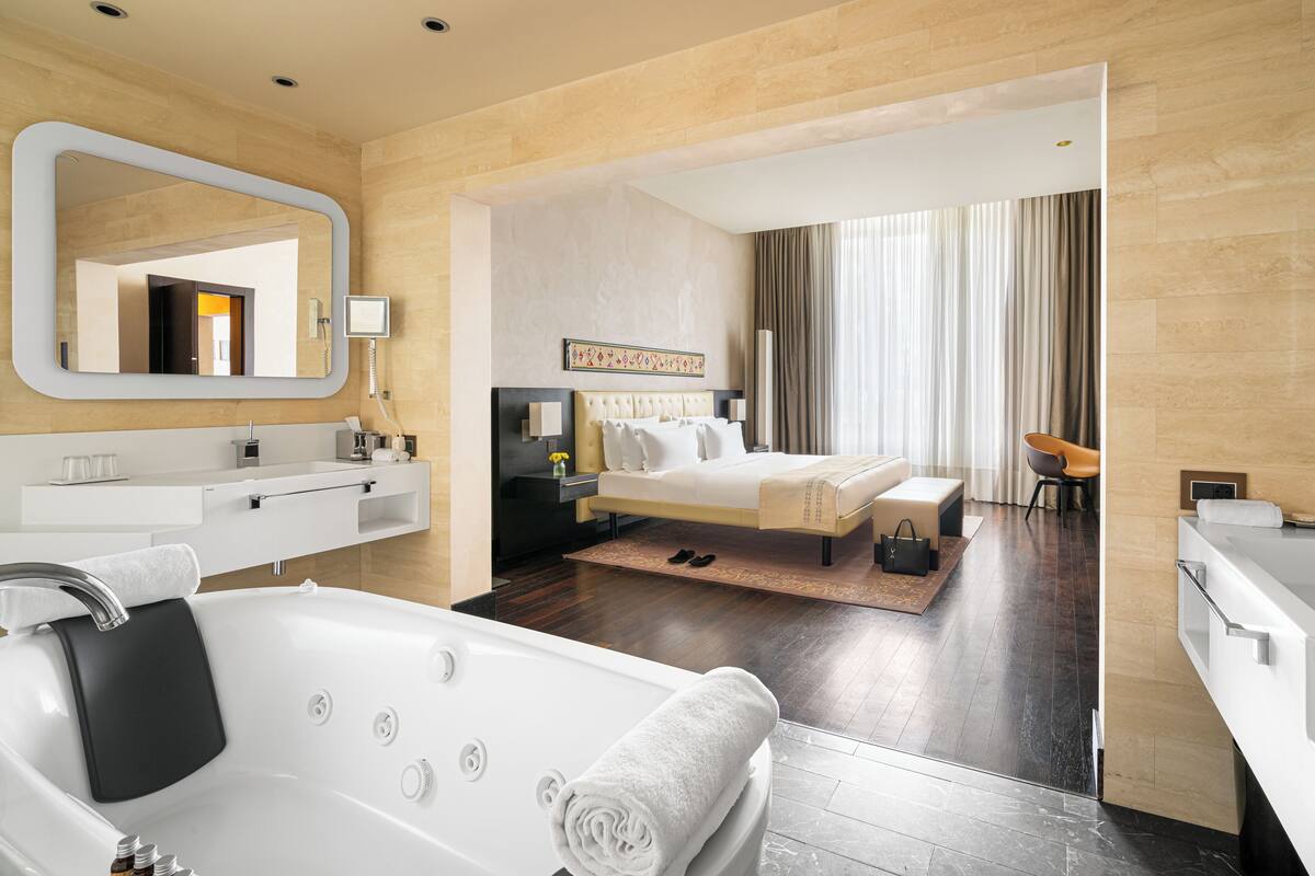 Una habitación de hotel moderna con una bañera grande, una cama con cabecero, un rincón de descanso con una silla y un espejo amplio.