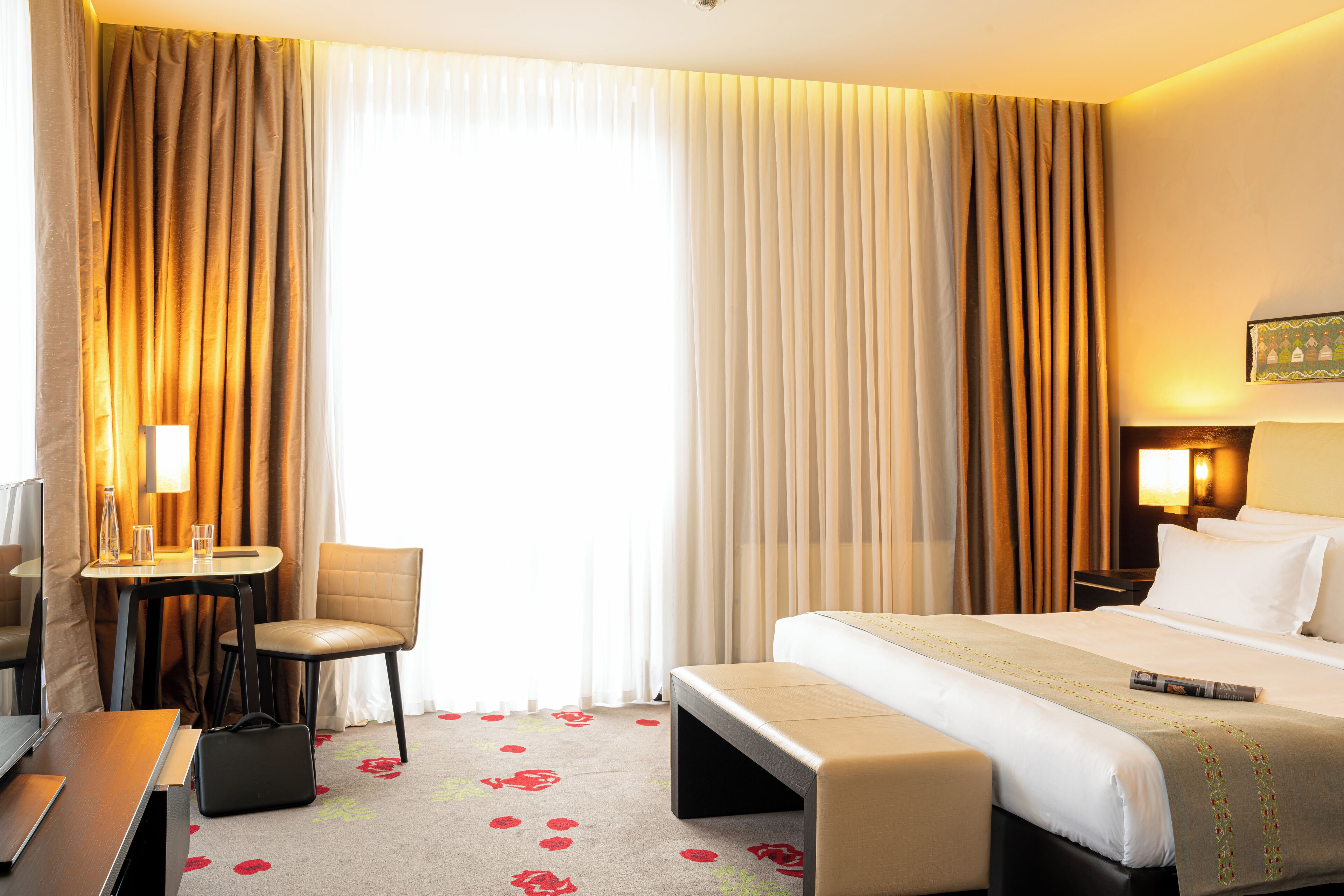 A hotel room with a bed, a desk, a chair, a bench, and a TV.