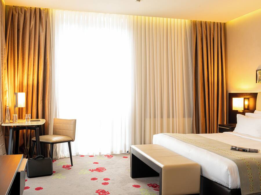 A hotel room with a bed, a desk, a chair, a bench, and a TV.