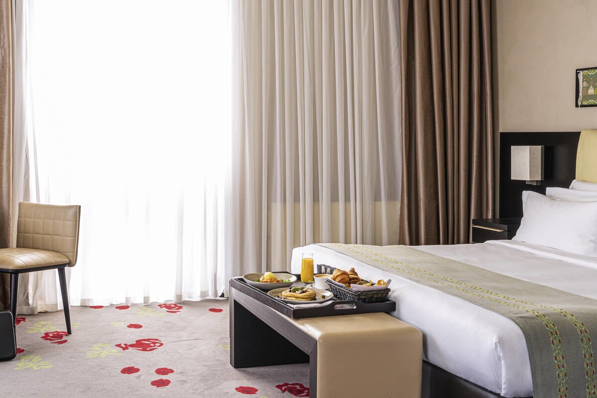 A hotel room with a bed, a bedside table with breakfast items, a chair, and a large window with curtains.
