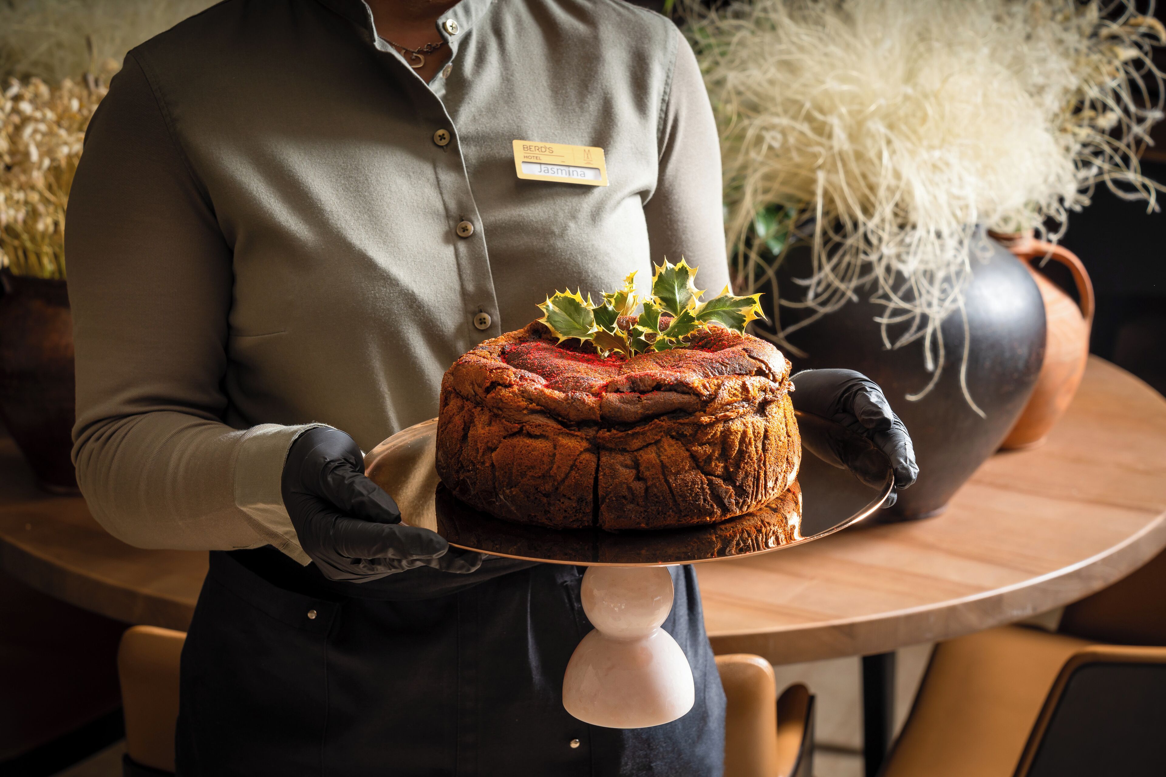 A person holding a large, round cake with a name tag, in a room with a wooden table and potted plants.