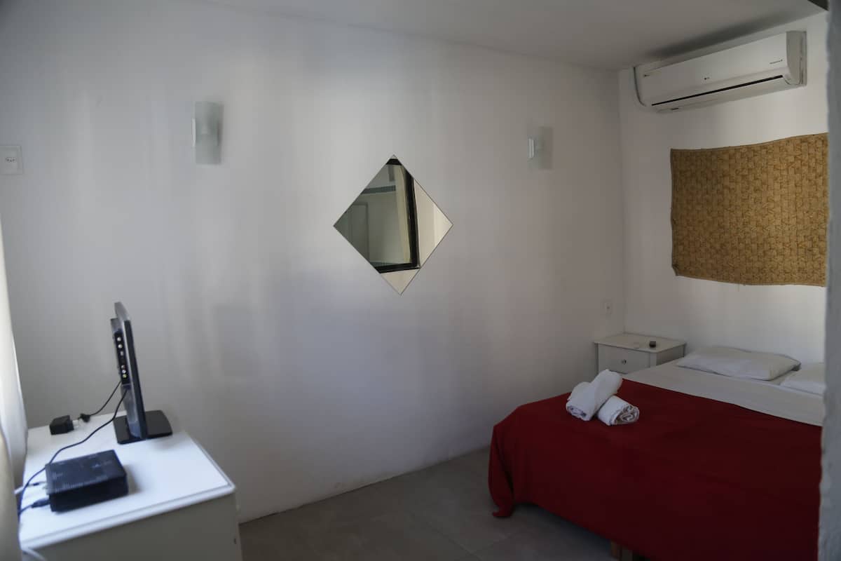 Una habitación pequeña y limpia con una cama, un escritorio con una computadora, un espejo y aire acondicionado.