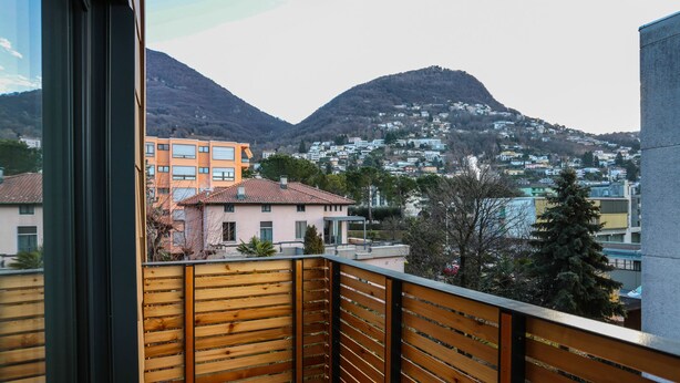 Un balcón con baranda de madera que da a una zona residencial con casas y montañas al fondo.