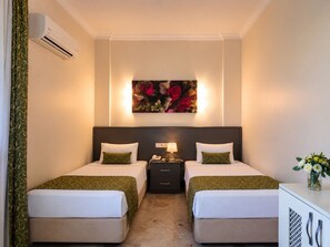 Een hotelkamer met twee bedden, een nachtkastje met een lamp, een airconditioning en een bloemig kunstwerk boven de bedden.