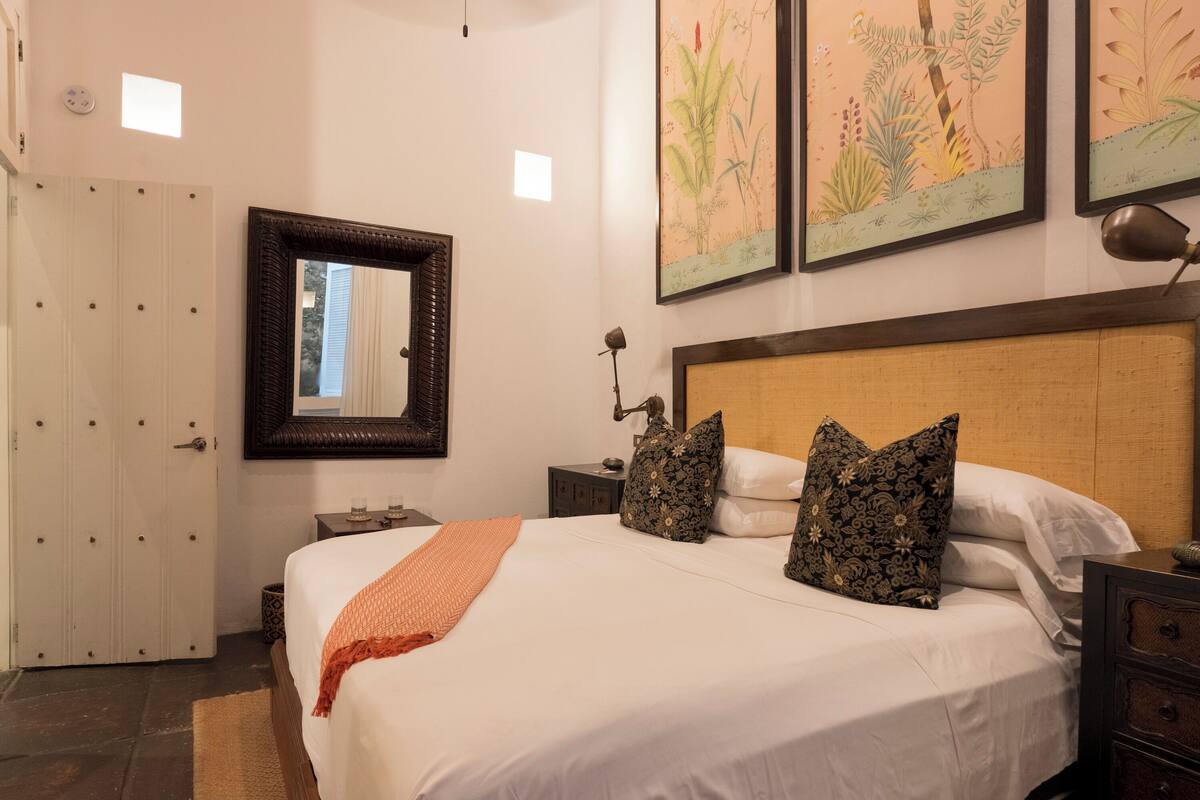 Una cama bien hecha con edredón blanco y manta naranja, cabecera de madera, dos mesitas de noche con lámparas, un espejo y dos impresiones enmarcadas con motivos botánicos en la pared.