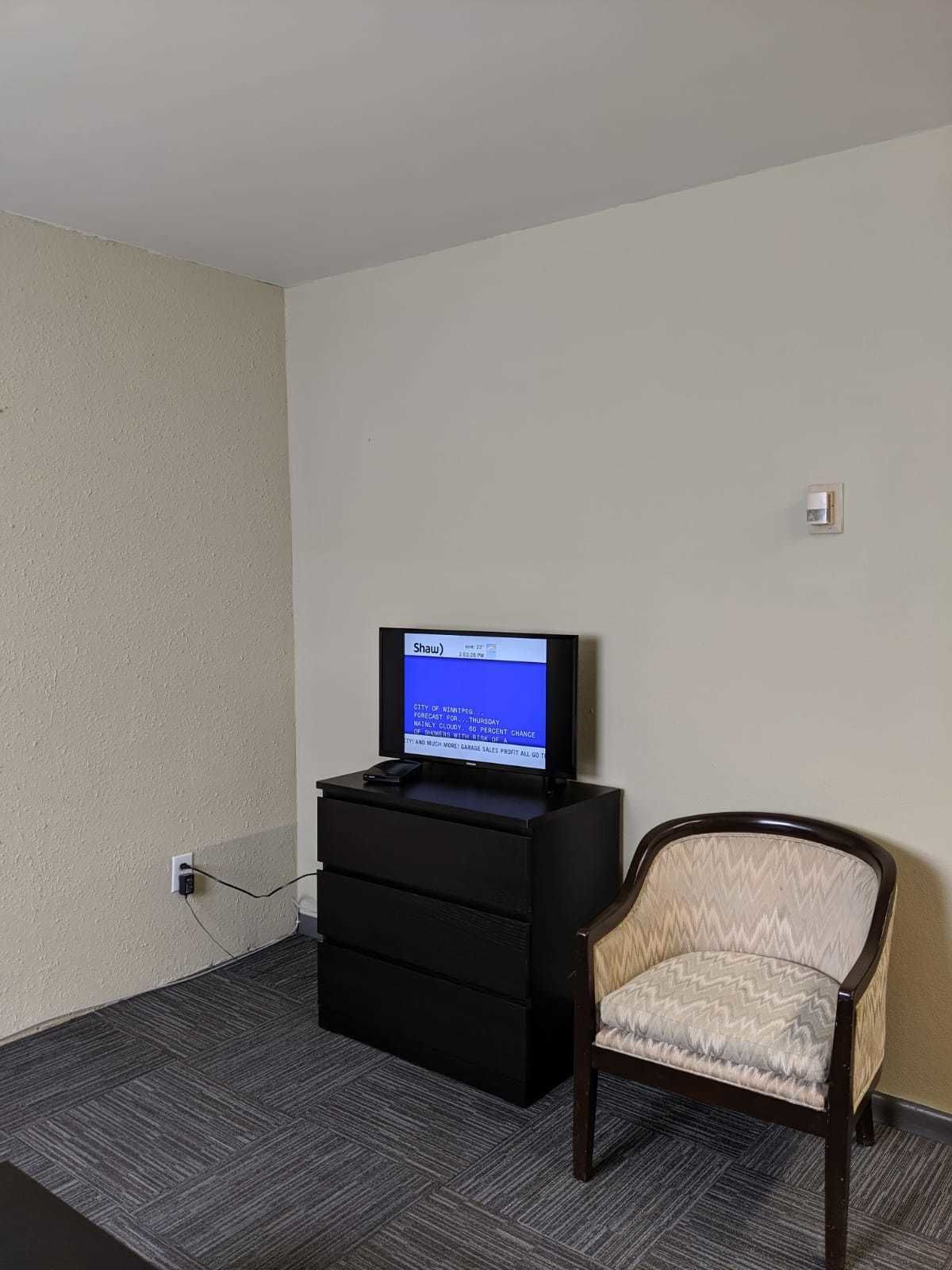 A hotel room with a television on a black stand, a wooden chair, and a carpeted floor.