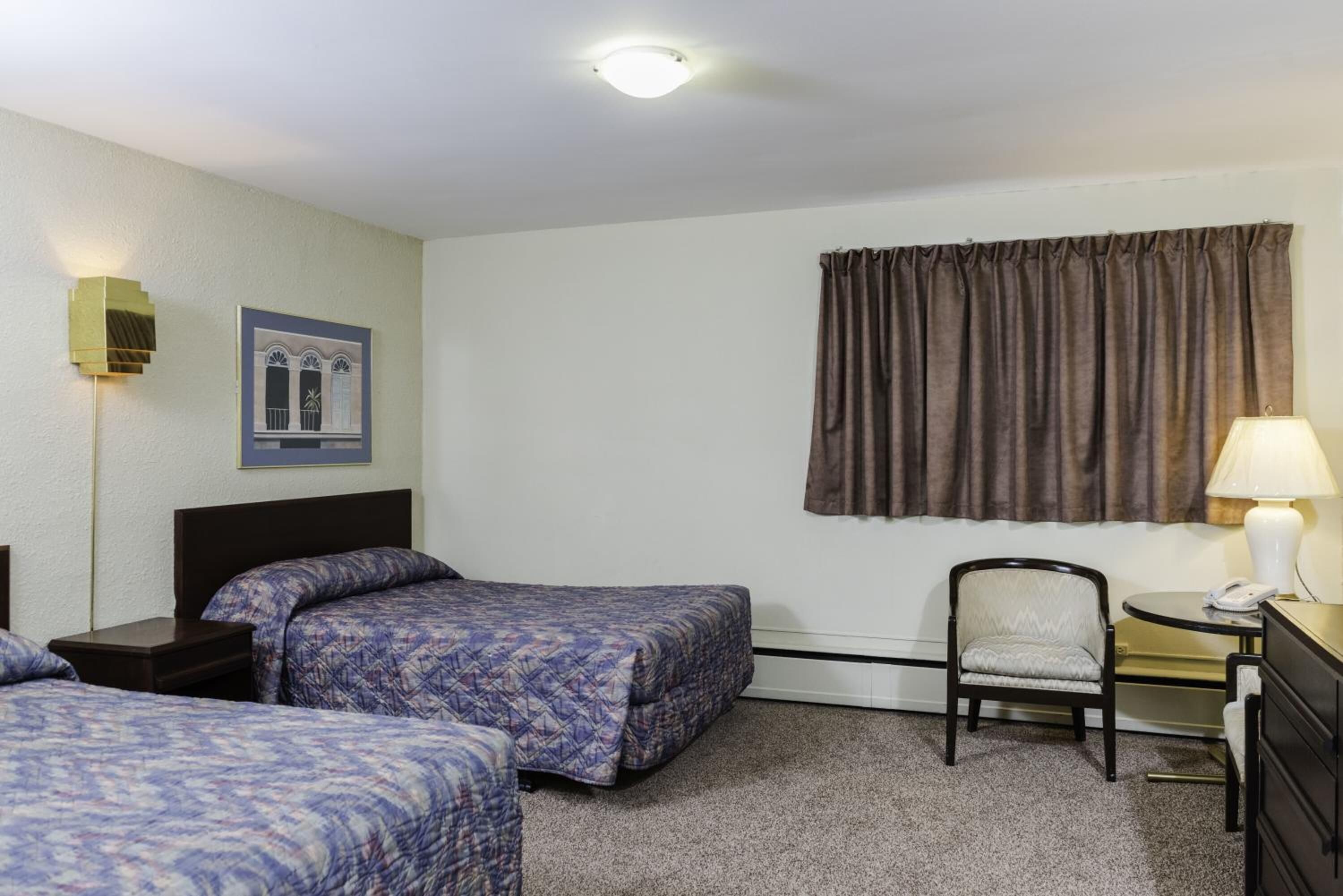 Habitación de hotel con dos camas, una silla, una mesita con lámpara, un cuadro en la pared y un ventanal con cortinas.