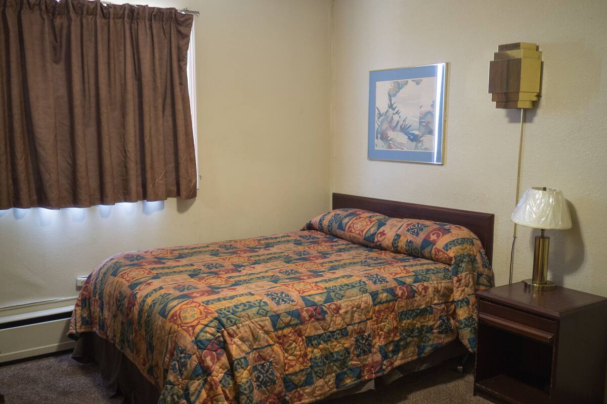 Un dormitorio con cama, mesita de noche, lámpara, ventana con cortinas y un cuadro en la pared.