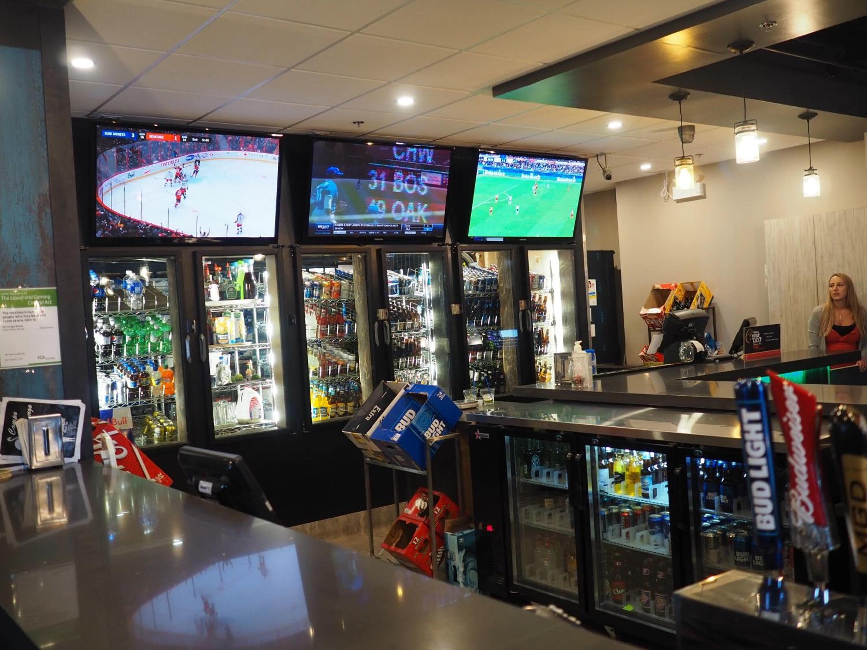 A bar area with multiple screens displaying sports events, a refrigerated display case with beverages, and a counter with a Bud Light sign.