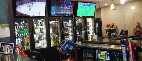 A bar area with multiple screens displaying sports events, a refrigerated display case with beverages, and a counter with a Bud Light sign.