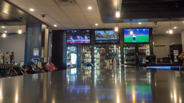 A bar area with multiple screens displaying sports events, a well-lit ceiling with hanging lights, and a polished floor. - The New Cavalier Inn (Winnipeg)