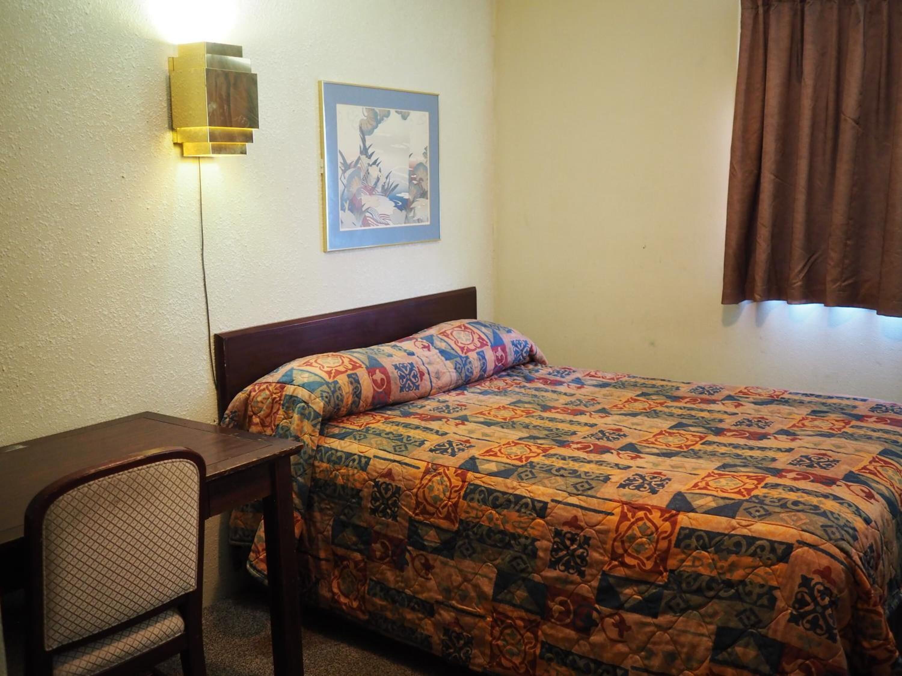 A bedroom with a bed, a desk, a chair, a wall-mounted lamp, and a framed picture.