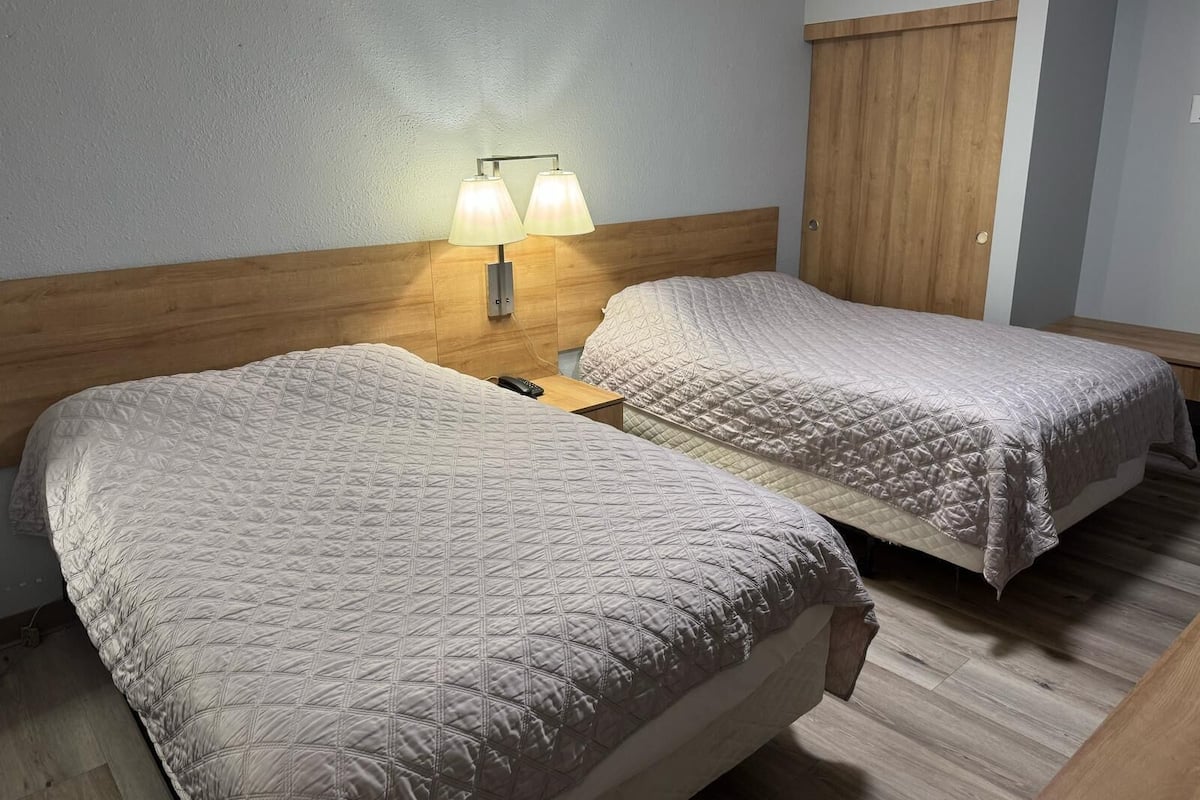 Habitación de hotel con dos camas, cabecero de madera, mesita de noche con lámpara y puerta de madera.