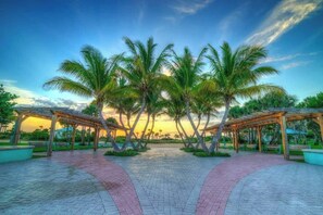 A paved walkway with palm trees and wooden pergolas, leading towards a sunset.